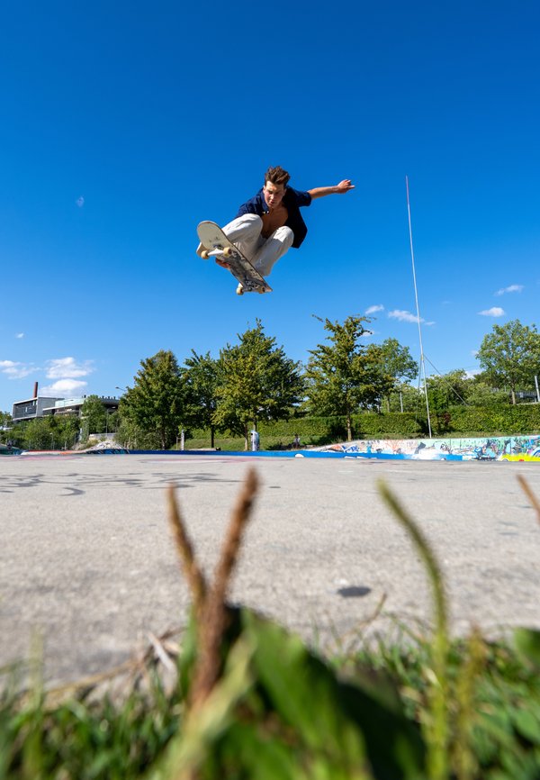 Ung mand iført hvide bukser og åben skjorte udfører et højt skateboardhop i en udendørs skatepark på en klar solskinsdag.