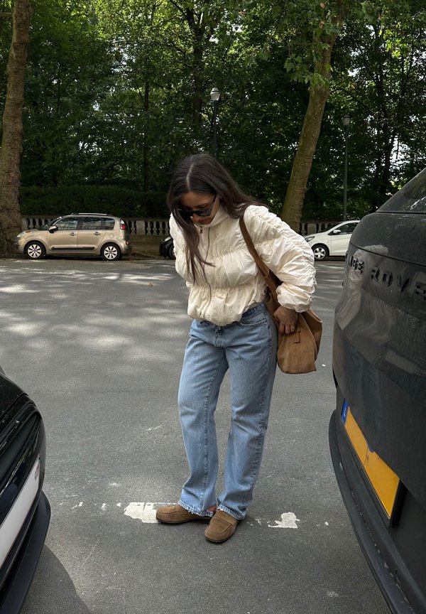 Joven mujer con gafas de sol, chaqueta crema, vaqueros azules y zapatos marrones, de pie entre coches aparcados en una calle arbolada.