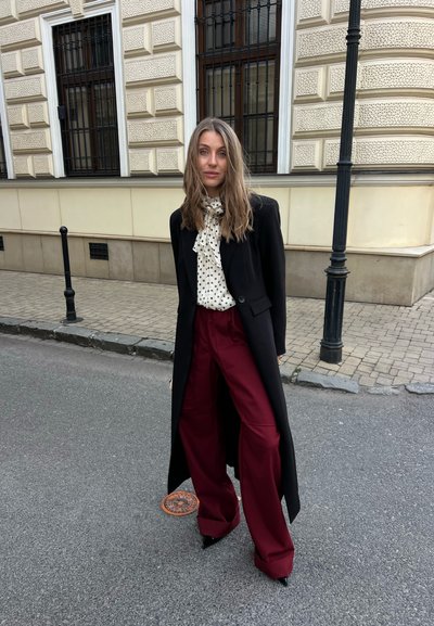 Mujer de pie en la calle de la ciudad con abrigo largo negro, blusa blanca con lunares y pantalones anchos de color burdeos cerca de un edificio beige.