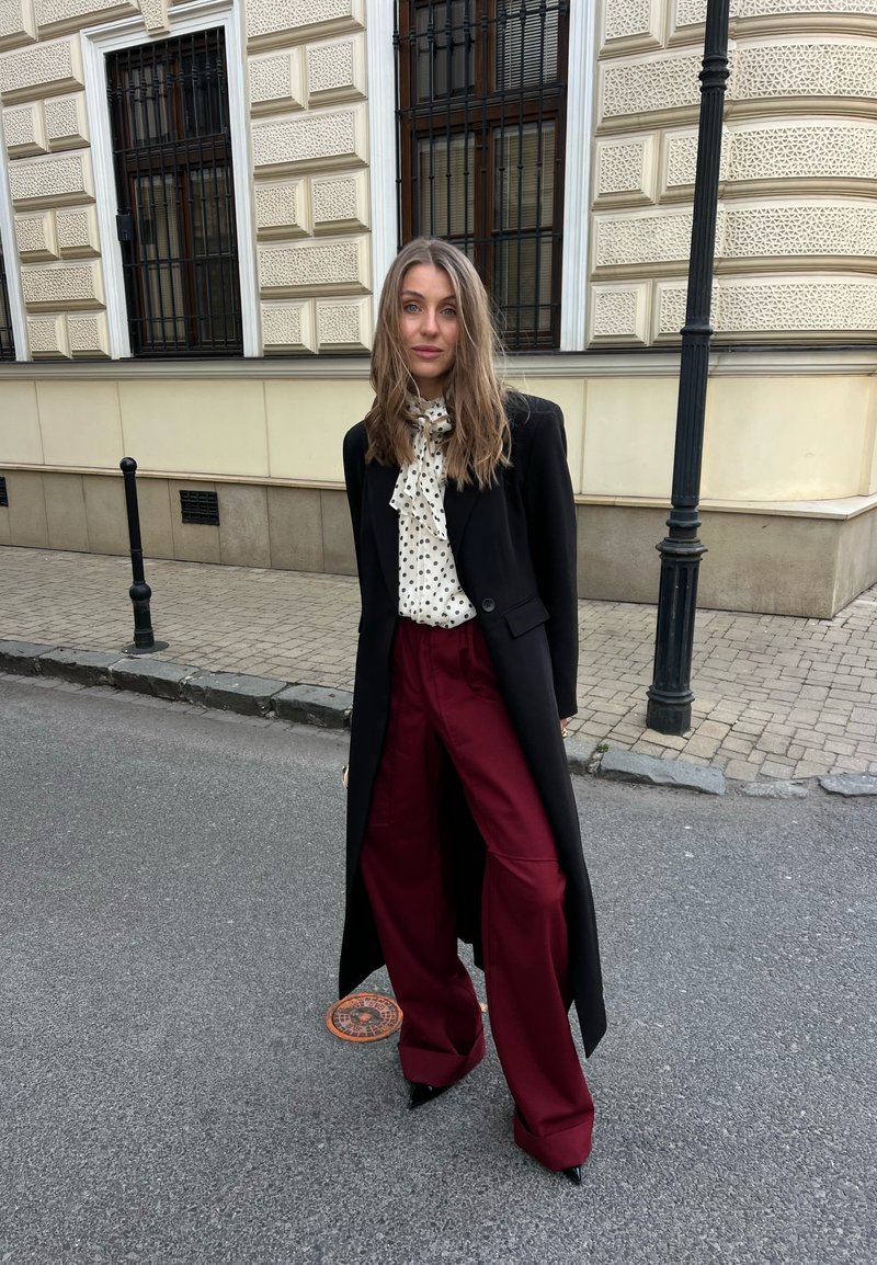 Femme debout dans une rue de la ville portant un long manteau noir, un chemisier blanc à pois et un pantalon large bordeaux près d'un bâtiment beige.