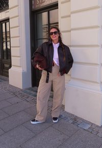 Rjava usnjena kratka jakna, bela srajca z gumbi, burgundska majica, bež cargo hlače, črne superge z belimi podplati, drži rjavo torbo.