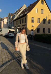 Jasne beżowe marynarki na białym topie i dopasowanej długiej spódnicy, w połączeniu z białymi trampkami. Sceneria przedstawia brukowane uliczki i żółty budynek.