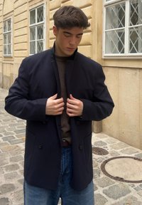 Marineblå blazer med høy krage, dobbelt knappelukking og sidelommer, kombinert med en brun genser og blå jeans. Glatt stofftekstur.