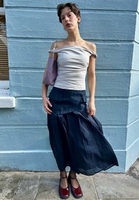 Μπεζ τοπάκι με ώμους έξω, ναυτικό μακρύ φούστα με πτυχώσεις και τσέπες, συνδυασμένο με κόκκινα παπούτσια και μωβ τσάντα ενάντια σε έναν ανοιχτό μπλε τοίχο.