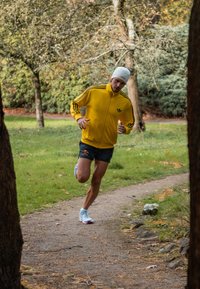 Żółta kurtka rozpinana z czarnymi paskami, niebieskie szorty i jasne buty do biegania. Biegacz znajduje się na ścieżce gruntowej wśród zielonej trawy i drzew.