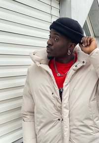 Light beige puffer jacket with a high collar, silver zip, and visible stitching; layered over a red t-shirt, completed with a black knitted beanie.