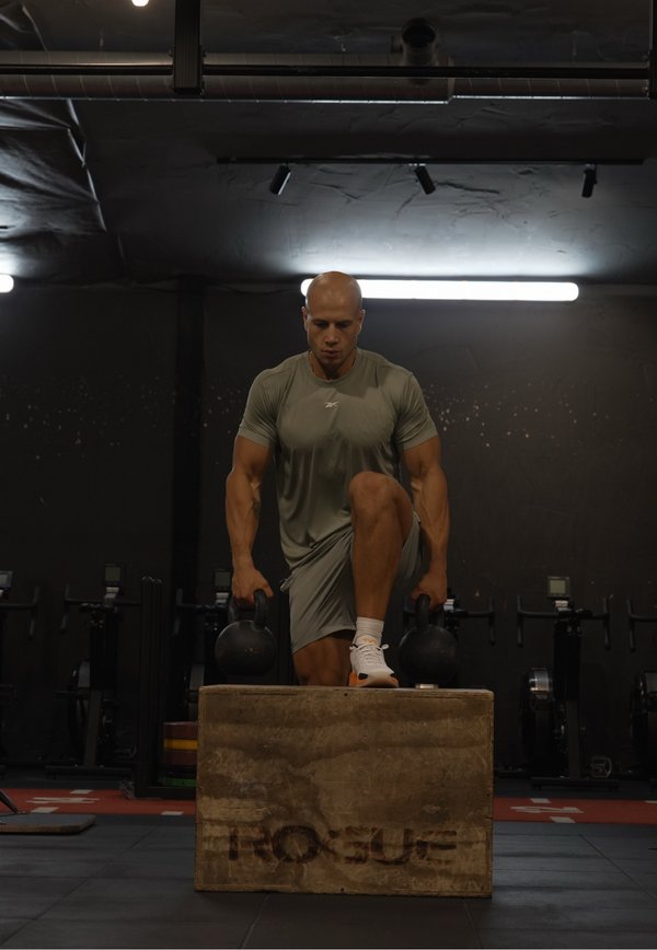 Homme en tenue de sport grise monte sur une boîte en bois en tenant des kettlebells dans une salle de sport faiblement éclairée avec du matériel d’exercice en arrière-plan.