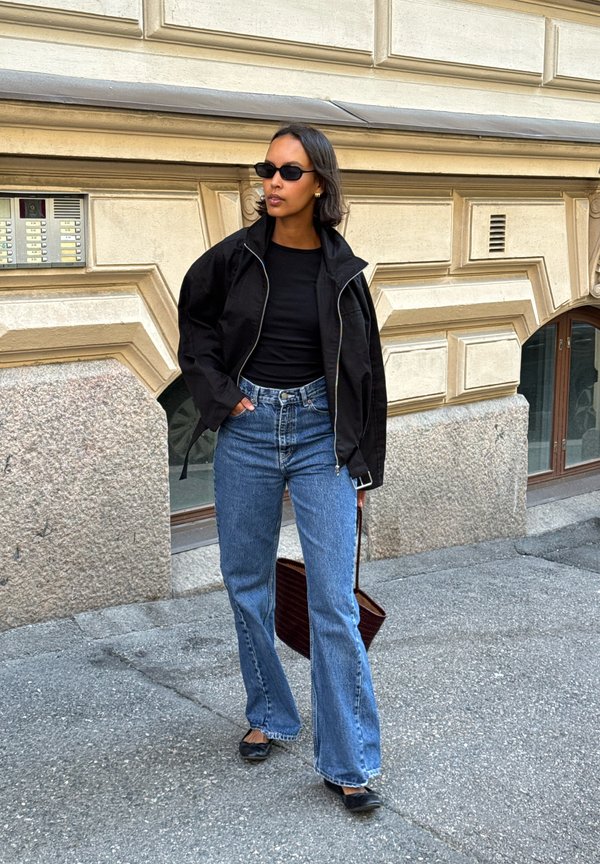 Mujer vestida con chaqueta negra, top negro, vaqueros azules, zapatos negros y gafas de sol, de pie en una calle con un edificio de piedra texturizada detrás.