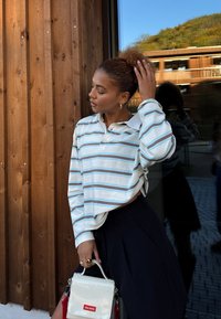 Stripete langermet poloskjorte i lyseblå med brune striper, kombinert med svarte bukser med høyt liv. En hvit blank veske med røde detaljer.