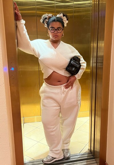 Suéter blanco texturizado, pantalones de chándal blancos y zapatillas plateadas, complementados con un bolso cruzado negro, de pie en un ascensor.