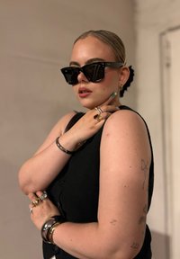 Top preto sem mangas com detalhes em botão, grandes óculos de sol pretos, múltiplos anéis e pulseiras, com um coque alto e tatuagens visíveis.