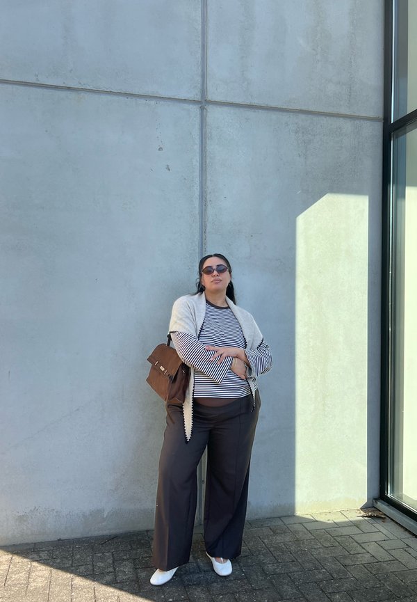 Mujer con gafas de sol, camisa de rayas, chal blanco y pantalones negros, de pie con los brazos cruzados contra una pared de hormigón cerca de una ventana de cristal.