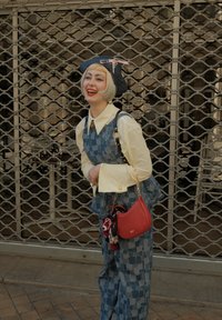 Persona con un corte de pelo bob rubio claro, vestida con un overol azul con estampado, blusa crema, sombrero negro con un lazo rosa y llevando un bolso rojo, sonriendo al aire libre.