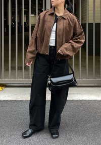 Hnědá semišová krátká bunda, bílý top, černé kalhoty s širokými nohavicemi a černá kabelka. Ploché černé boty, na bundě je viditelný kovový kovový detail.