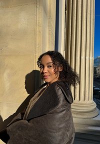 Vêtement extérieur texturé brun foncé avec un col en fourrure. Le tissu semble doux, avec une coupe ample, se détachant sur un fond en pierre claire.