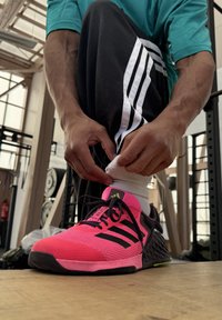 Jasnoróżowy but sportowy z czarnymi akcentami i teksturowanym materiałem, zestawiony z białą skarpetą. W tle znajduje się sprzęt do ćwiczeń i drewniana podłoga.