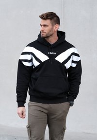 Črna jopa s kapuco z belimi diagonalnimi črtami čez prsa in rokave. Ima sprednji žep in nastavljivo kapuco z vrvicami.