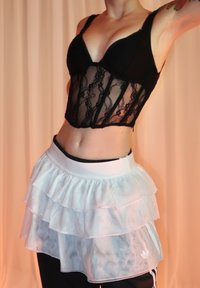 Bralette en dentelle noire à design structuré, associée à une jupe blanche transparente à volants. La jupe présente une ceinture élastique et un branding subtil.