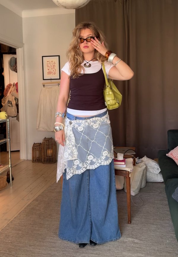 Denim maxi sukně s krajkovým překrytím, přiléhavý černý top, bílý tričko pod ním, zelená kabelka, vrstvené náramky a černé boty.