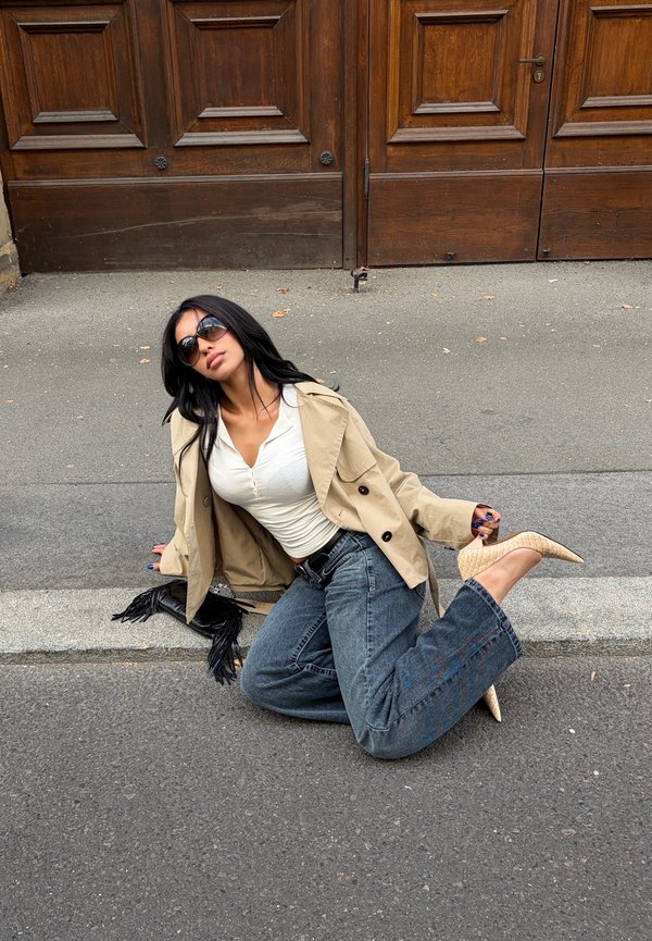 Una mujer con gafas de sol, una chaqueta beige, un top blanco y vaqueros posa en una acera frente a una gran puerta de madera, con un bolso beige en la mano.