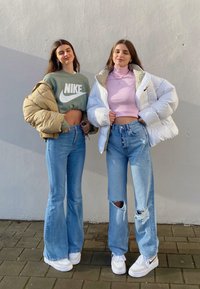 Dwie kobiety noszą casualowe stroje: jedna w zielonym, krótkim dresie Nike i beżowej kurtce puchowej, druga w różowej bluzce i białej kurtce puchowej. Obie w niebieskich dżinsach.