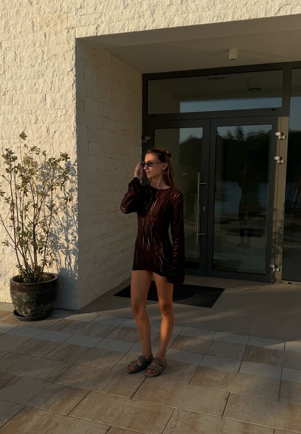 Femme portant des lunettes de soleil et une robe sombre debout devant un bâtiment avec des portes en verre près d'une plante en pot à la lumière tardive de l'après-midi.