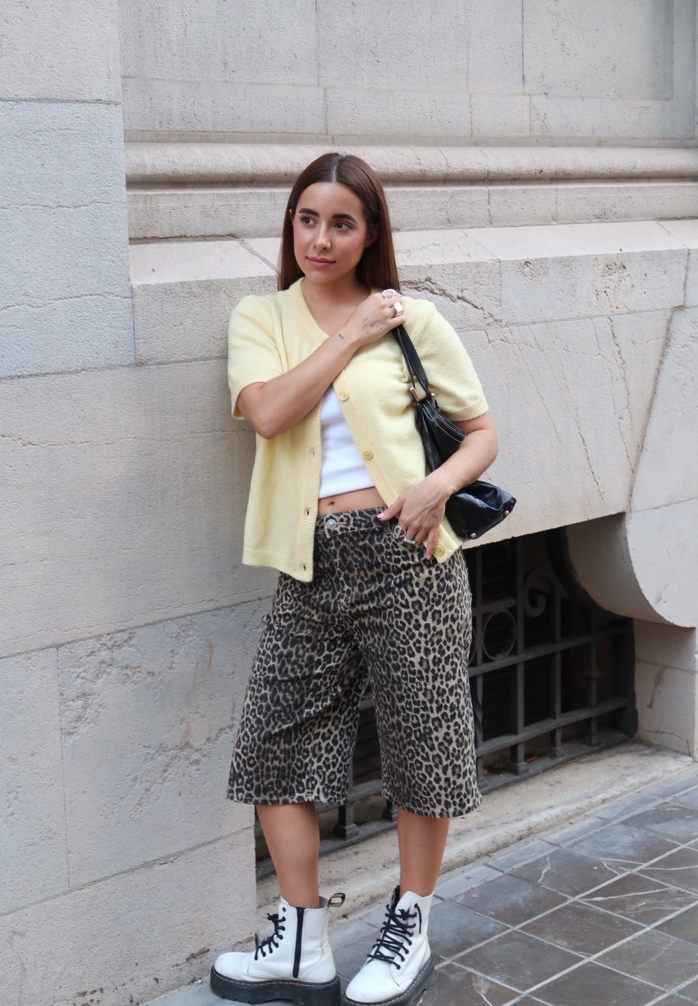 Svijetložuti kratki kardigan s kratkim rukavima, bijeli crop top, hlače širokog kroja s uzorkom leopard, i bijele čizme na vezanje s crnim potplatima. Crna torba.
