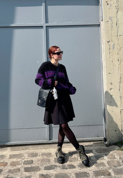 Suéter de lana peluda a rayas negro y morado, falda negra acampanada, pantys negros, botas de combate verdes, bolso de hombro negro con charm, gafas de sol.