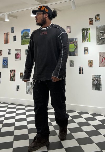 Camisa negra de manga larga con costuras grises, pantalones negros y zapatillas marrones. Los accesorios incluyen una gorra negra y una cadena plateada. Suelo de cuadros.