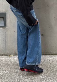 Laza szabású kék farmernadrág szálazott szegéllyel, fekete sportcipővel párosítva, amely piros részletekkel és texturált talppal rendelkezik.