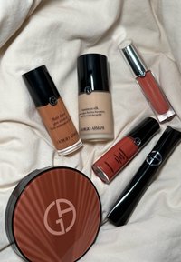 Six produits de maquillage Giorgio Armani, y compris des fonds de teint liquide, des brillants à lèvres, du mascara et une poudre compacte, disposés sur un fond en tissu crème.