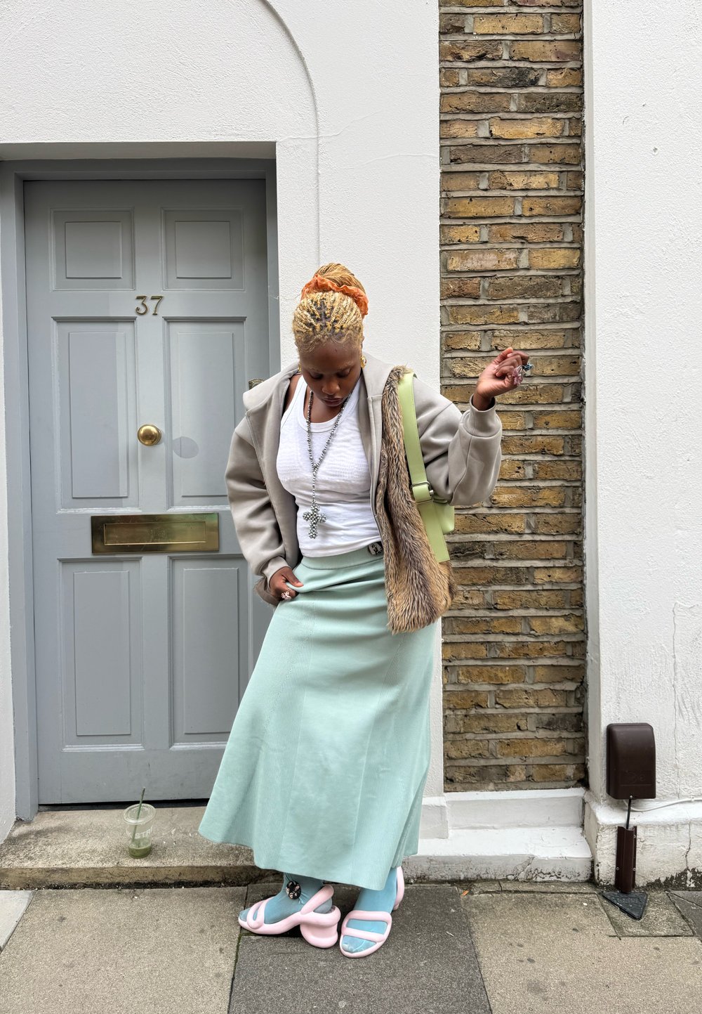 Egy személy bézs kapucnis pulóverben műszőrmével, fehér felsővel és menta zöld szoknyával. Buborékrágó rózsaszín papucsot visel, és italt tart a kezében. Kőfal a háttérben.