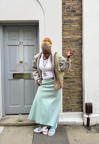 Egy személy bézs kapucnis pulóverben műszőrmével, fehér felsővel és menta zöld szoknyával. Buborékrágó rózsaszín papucsot visel, és italt tart a kezében. Kőfal a háttérben.