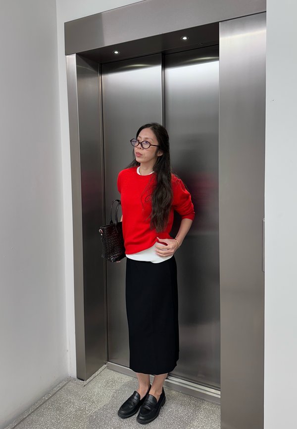 Una mujer con gafas, un jersey rojo y una falda negra está de pie frente a las puertas cerradas de un ascensor de acero inoxidable.