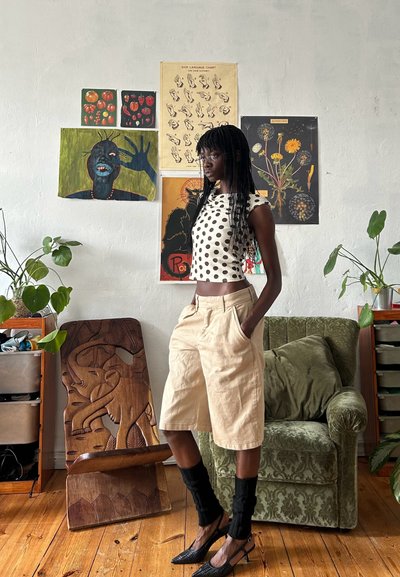 Pantalones cortos anchos beige combinados con un top recortado blanco con puntos negros. Zapatos de tacón altos negros y calentadores de piernas texturizados negros. Tallado de elefante de madera cerca.