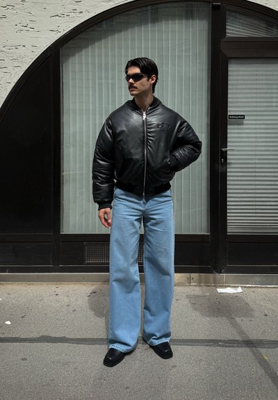 Chaqueta de bombardero de cuero negra con puños acanalados, combinada con pantalones vaqueros de pierna ancha de color azul claro. El modelo lleva gafas de sol negras y zapatos negros.