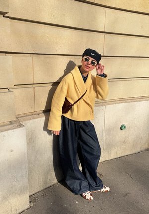 Persona con un abrigo amarillo, pantalones anchos oscuros, zapatos con estampado de vaca, gafas de sol blancas y gorra negra de Nike, apoyada contra una pared de piedra beige bajo la luz del sol.