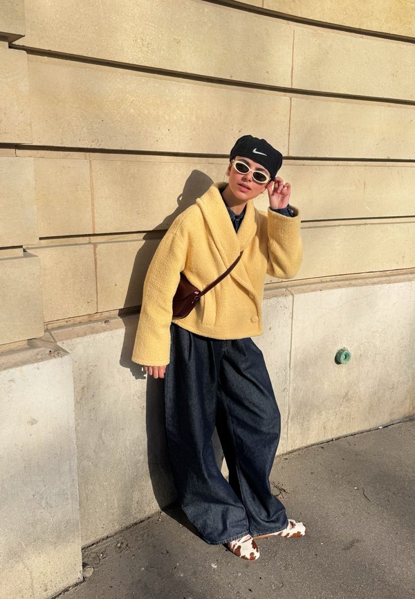 Άτομο που φοράει κίτρινο παλτό, φαρδιά σκουρόχρωμα παντελόνια, παπούτσια με σχέδιο αγελάδας, λευκά γυαλιά ηλίου και μαύρο καπέλο Nike, στηρίζεται σε έναν μπεζ πέτρινο τοίχο στο ηλιακό φως.