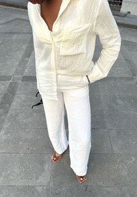 Camicia bianca con texture, a maniche lunghe e tasca, abbinata a pantaloni bianchi larghi. Persona che indossa sandali con tacco metallici su una superficie pavimentata.