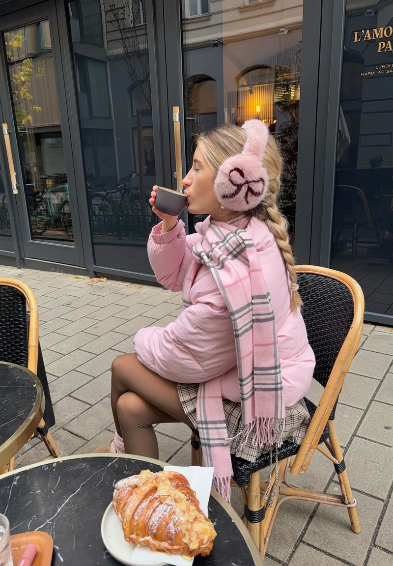 Pink puffer jacket, checkered scarf, and fluffy earmuffs. Holding a black cup, seated at a table with a croissant on a white plate.