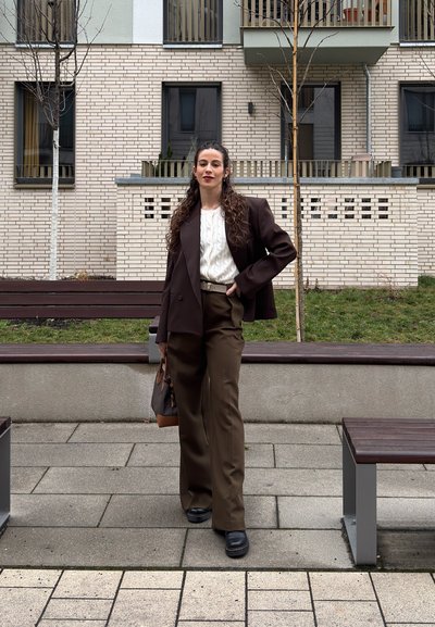 Mujer con cabello largo y rizado de pie al aire libre, vistiendo traje marrón, suéter blanco, zapatos negros, sosteniendo un bolso cerca de un edificio de ladrillos beige con árboles sin hojas.