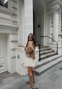 Beige cropped jakke, hvid blondekjole, brune knæhøje støvler. Langt hår, solbriller. Trapper og søjler fra en bygning i baggrunden.