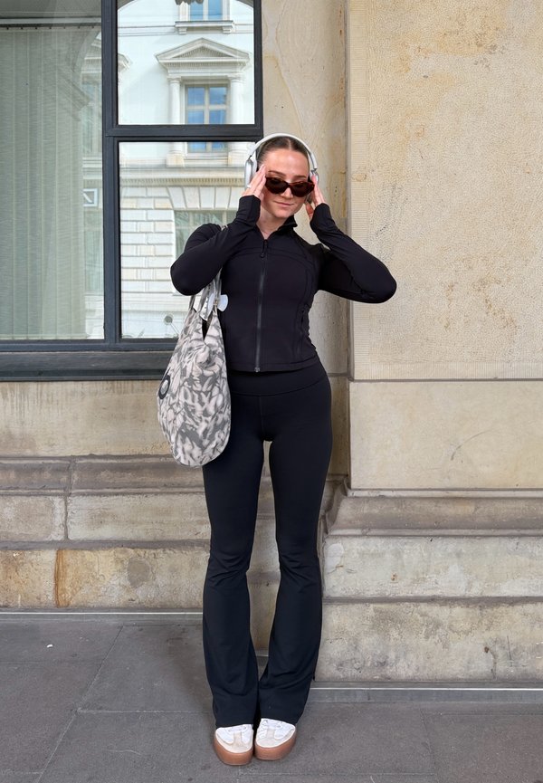 Persona vestida con ropa deportiva negra, auriculares blancos, gafas de sol y zapatos de plataforma blancos, está junto a una pared de piedra sosteniendo una bolsa grande con estampado.
