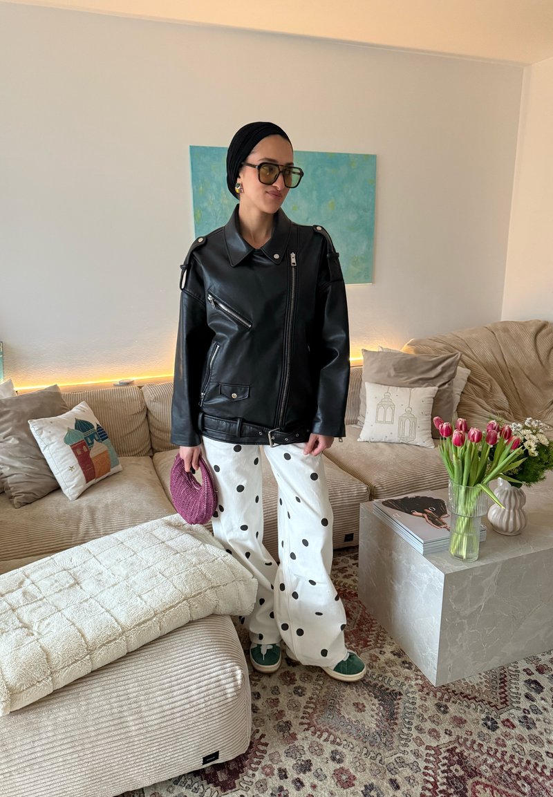 Femme en veste en cuir noire et pantalon blanc à pois tenant un sac violet, debout dans un salon moderne avec un canapé beige et des tulipes.