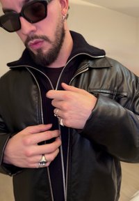 Homme barbu avec des lunettes de soleil en train de fermer la fermeture éclair d'une veste en cuir noire, portant plusieurs bagues en argent et des boucles d'oreilles, à l'intérieur.