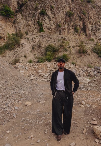 Hombre con chaqueta negra, camisa blanca y pantalones negros anchos de pie sobre terreno rocoso con una colina empinada y rocosa detrás de él.