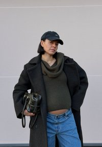 Personne enceinte portant un manteau sombre, un pull côtelé vert olive, un jean bleu clair et une casquette noire. Elle tient un petit sac à main noir.