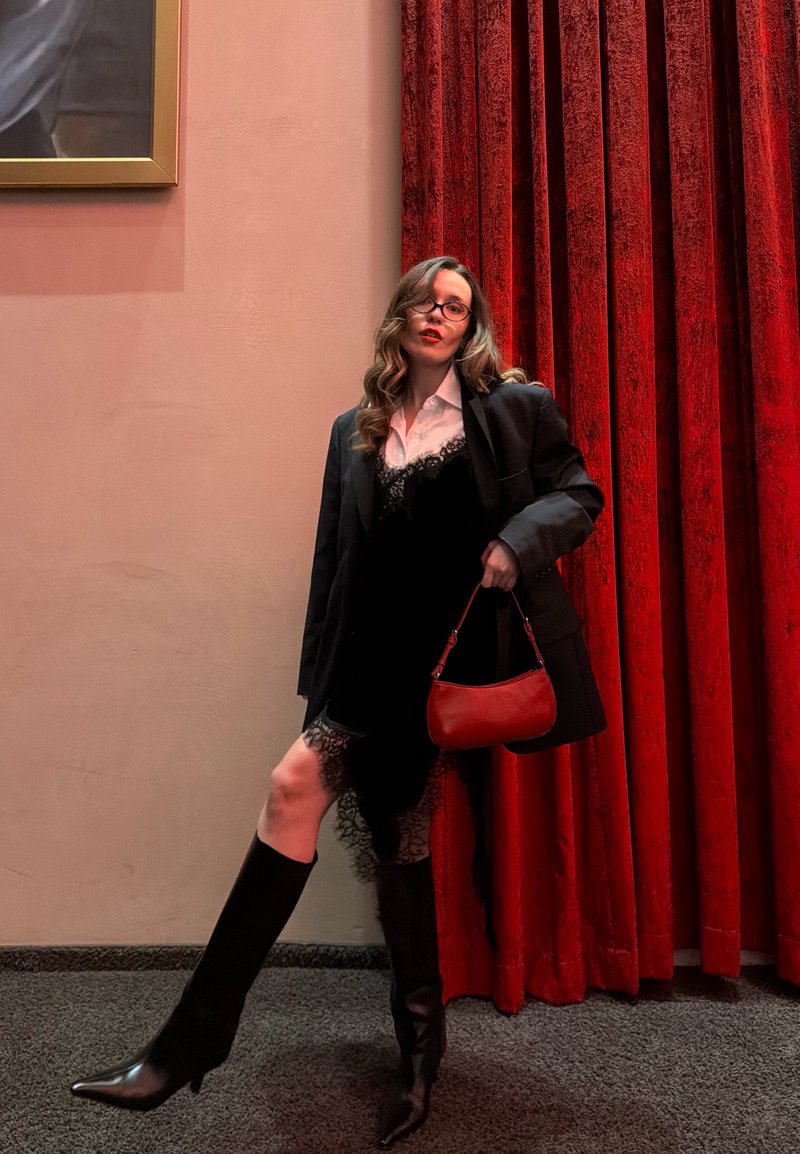 Femme aux cheveux ondulés portant des lunettes, une robe en dentelle noire, un blazer noir et des bottes montantes, posant avec un sac à main rouge devant des rideaux rouges.