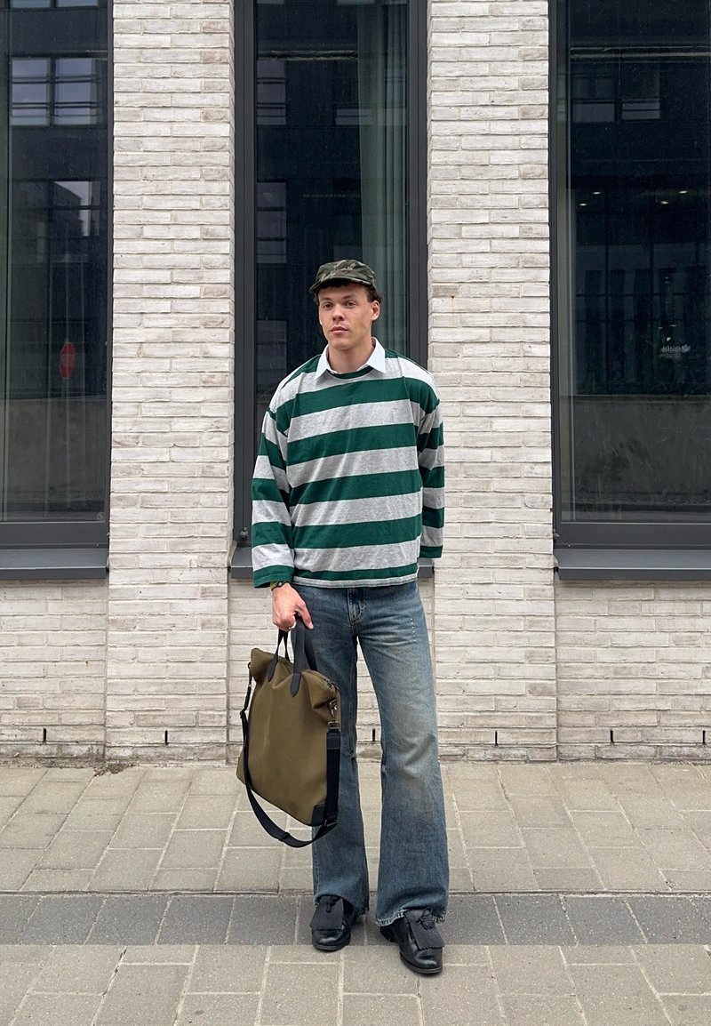 Young man wearing green and grey striped jumper and cap, holding a green tote bag, standing on a city sidewalk in front of a brick wall.