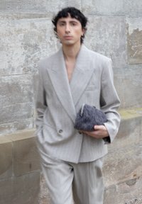 Persona joven caminando junto a un muro de piedra, vestida con un traje gris claro, sosteniendo un pequeño bolso de mano texturizado de color gris oscuro en una mano.